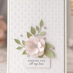 May include: A rectangular greeting card with a white embossed basketweave pattern background. The card features a layered pale pink paper flower with a central crystal, sage green paper leaves, and small clear rhinestones. A white label reads "SENDING YOU all my love" in black text.
