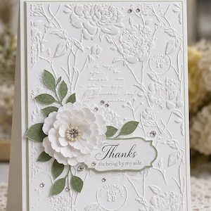 Puede incluir: Una tarjeta de felicitación blanca con un patrón floral en relieve, una flor de papel en capas con un cristal central y hojas de papel verde. Incluye una etiqueta con el texto "Thanks for being by my side" decorada con pequeños brillantes.