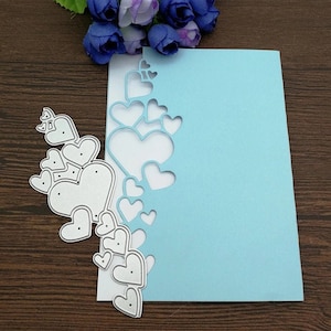 Puede incluir: Un troquel met&aacute;lico con una serie de corazones de varios tama&ntilde;os, dispuestos en el lado izquierdo de una tarjeta azul claro. El troquel crea aberturas en forma de coraz&oacute;n. Un racimo de flores azules est&aacute; en el fondo.