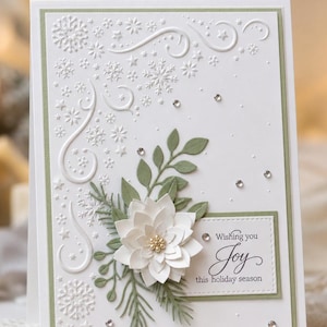 May include: A white greeting card with a sage green border featuring embossed snowflakes and swirls. A layered white paper flower with a gold beaded center sits atop sage green paper foliage. A rectangular label reads Wishing you Joy this holiday season.