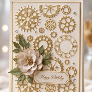 May include: A cream-coloured greeting card featuring gold embossed gear and clock motifs, a layered paper flower with green leaves, and a central rectangular label with the text Happy Birthday in elegant script, accented by small clear rhinestones.