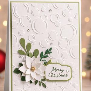 May include: A white greeting card featuring an embossed bubble pattern, a layered white poinsettia with gold centers, green paper foliage, and a rectangular label with the text Merry Christmas, accented by small clear adhesive rhinestones.