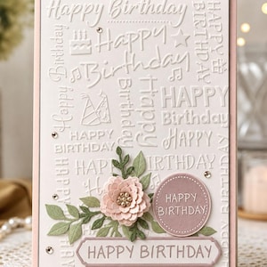 Op de afbeelding: Een rechthoekige wenskaart met een lichtroze rand en een witte reliëfachtergrond met de tekst Happy Birthday, feesthoedjes en taarten. Het is versierd met een gelaagde papieren bloem, groene bladeren en een rond label met tekst.