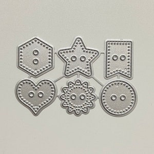 Peut inclure: Un ensemble de six formes découpées au die en métal, chacune avec un motif de bouton. Les formes comprennent un cœur, une étoile, un cercle, un hexagone, une fleur et une bannière.