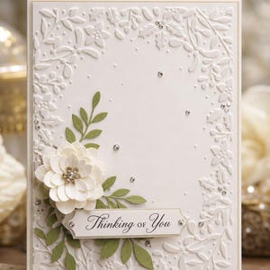 Pode incluir: Um cartão de felicitações retangular com uma borda floral branca em relevo, apresentando uma flor de papel branca em camadas com um centro de cristal, folhas de papel verdes, pequenos strass transparentes espalhados e uma etiqueta com o texto Thinking of You.