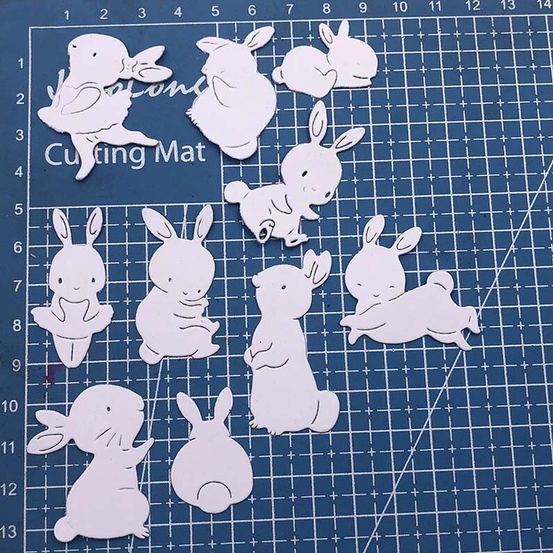 May include: Nine white die-cut bunny rabbit shapes on a blue cutting mat. The bunnies are in various poses, including standing, sitting, and lying down. The bunnies are all facing forward.