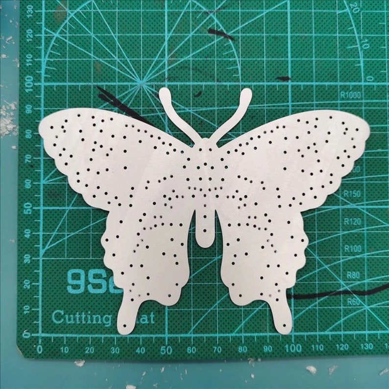 Może przedstawiać: Biały papierowy wycinany motyl z kropkowanym wzorem. Motyl znajduje się na zielonej macie do cięcia z białymi oznaczeniami.