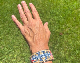 May include: A colorful beaded bracelet with the letters "BHF" in blue and red, set against a tan background. The bracelet has a silver-colored metal band. The image is taken outdoors on a sunny day.