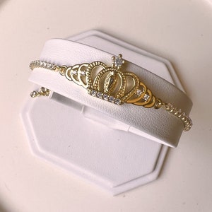 May include: A gold bracelet with a crown charm and clear cubic zirconia stones. The crown features a small image of the Virgin Mary.