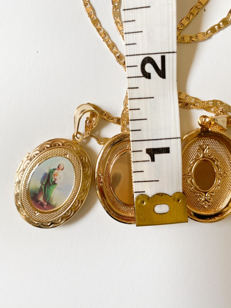 Puede incluir: Collar de cadena de oro con dos colgantes ovalados. Un colgante presenta una imagen religiosa de un hombre con ropa verde. El otro colgante est&aacute; vac&iacute;o.