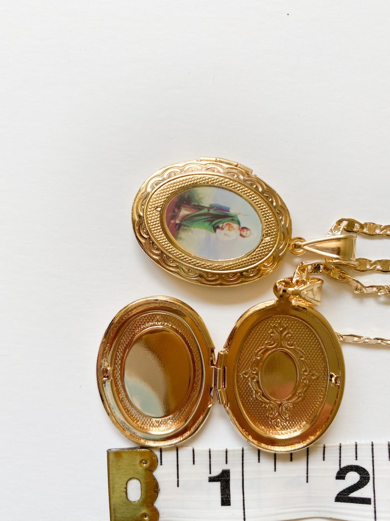 Puede incluir: Collar con dije ovalado de oro, con una imagen religiosa en la parte delantera y un dise&ntilde;o decorativo en la parte posterior. El dije est&aacute; abierto y la cadena es visible.
