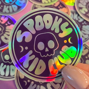 May include: A holographic sticker with the text "Spooky Kid Club" in a rainbow iridescent color scheme. The sticker features a skull with a crescent moon and star on either side.