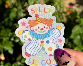 May include: A holographic sticker featuring a cartoon clown with a red nose, a yellow and orange hat, and a blue and white striped shirt. The sticker says "Silly Lil Guy" in rainbow letters.
