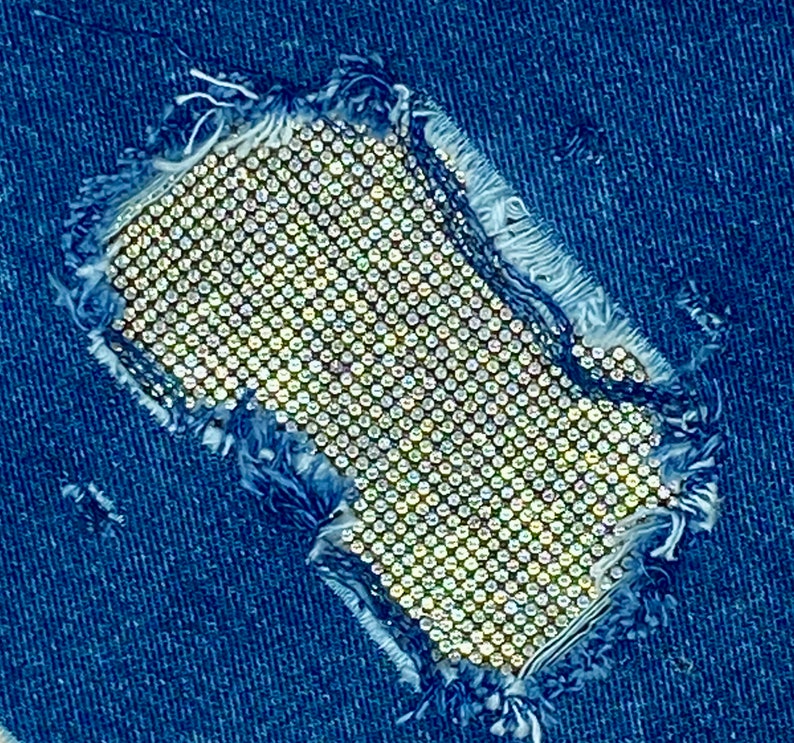 May include: A close-up of a denim fabric with a hole that is covered with a sparkly, silver-toned material. The material is made up of small, round rhinestones.