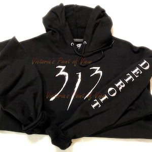 Puede incluir: Sudadera con capucha negra con un gráfico blanco del número 313 y la palabra "HORRHED" en la manga.