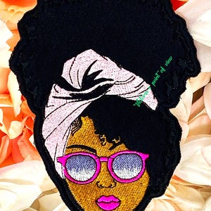 May include: Embroidered patch featuring a woman with a pink headscarf and pink sunglasses. Her hair is styled in an afro. The patch is black with a white outline.