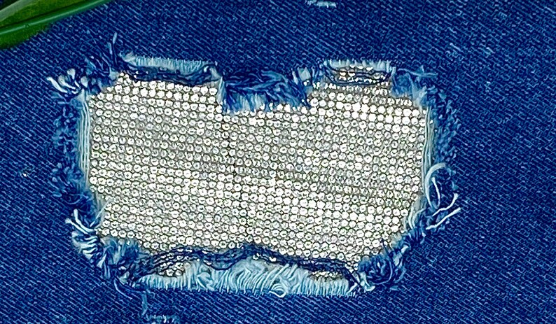 May include: A close-up of a blue denim fabric with a hole that has been patched with a silver sequined fabric.