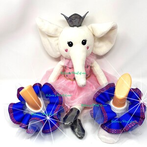 Puede incluir: Una muñeca de peluche de elefante blanco con un tutú rosa y una corona plateada. La muñeca lleva calcetines azules y blancos con un lazo de tul y cinta azul. El texto "Victoria's Point of view" es visible en los calcetines.
