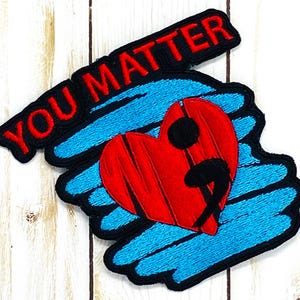 May include: Embroidered patch with a blue and black background. The patch features a red heart with a black semicolon inside. The text "YOU MATTER" is embroidered in red on the top of the patch.