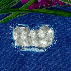 May include: A close-up of a blue denim fabric with a hole that has been patched with a silver sequined fabric.