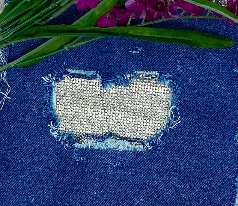 May include: A close-up of a blue denim fabric with a hole that has been patched with a silver sequined fabric.