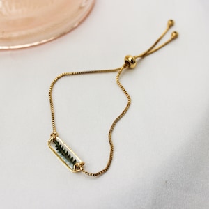May include: Gold-toned adjustable bracelet featuring a rectangular charm with a preserved fern leaf. The delicate chain and charm are set against a white background, with a blurred rose-gold colored object in the upper left corner.