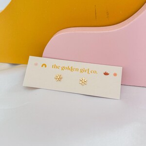 May include: A pair of gold-colored snowflake stud earrings on a cream-colored card with the text "the golden girl co." The card is placed on a pink and yellow background.