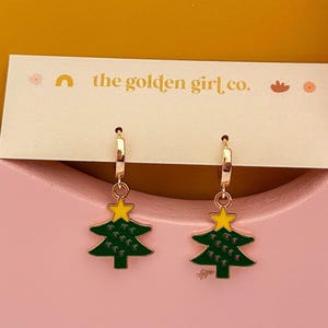 May include: Gold-coloured hoop earrings featuring green Christmas tree charms with yellow star toppers. The earrings are presented on a pink surface with a gold and white tag that reads "the golden girl co."
