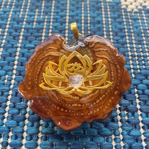 May include: A gold-colored pendant featuring a lotus flower with an Om symbol in the center. The pendant is set in a brown resin base with a wood-like texture.