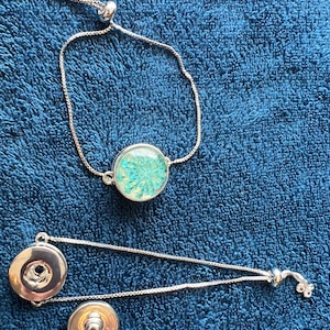 May include: Two silver adjustable bracelets with a floral design. One bracelet has a round, clear pendant with blue flowers inside. The other bracelet has two round, silver buttons.