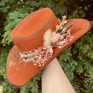 May include: An orange felt cowboy hat with a decorative floral ribbon and feather accents. The hat features a dark brown brim and is adorned with dried flowers and a fluffy accent. A unique, handcrafted accessory.