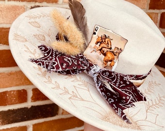 Puede incluir: Un sombrero de vaquero blanco con una pluma marrón y blanca, una bandana marrón y blanca y una carta de juego con una reina. La carta muestra a una mujer montando un caballo.