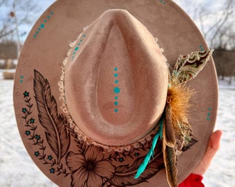 May include: A tan fedora-style hat with a wide brim, adorned with hand-painted floral and feather designs in brown and turquoise. The hat features a decorative feather and fabric accent on the side, adding a bohemian flair.