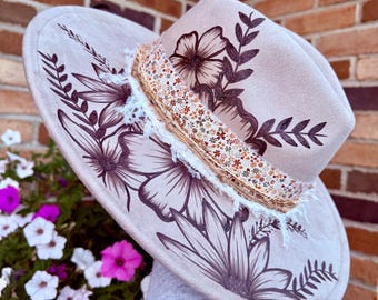 May include: A light beige fedora hat featuring dark brown floral line art illustrations on the crown and brim. The hat is accented with a layered band consisting of a floral patterned fabric strip, a frayed white lace trim, and twine.