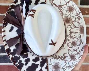 May include: A white cowboy hat with a decorative brim. The brim features a cow print, floral pattern, and brown accents. The hat is embellished with feathers and stitched details, creating a unique western-inspired accessory.