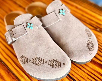 May include: A pair of light taupe suede clogs featuring a side strap with a metallic buckle and a small turquoise four-petal flower accent. The front of each shoe is decorated with a series of dark brown geometric patterns etched into the suede.