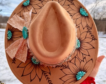 May include: A tan fedora-style hat with a wide brim, decorated with black sunflower outlines and turquoise-colored stones. A peach-colored fabric bow and lace trim adorn the hat. The hat is held up against a snowy background.