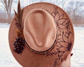 Puede incluir: Un sombrero fedora color canela con ala ancha, decorado con un diseño floral negro y plumas. El sombrero tiene una textura marrón similar al ante y una banda decorativa. El diámetro del ala es de aproximadamente 35.5 cm.