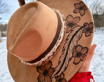 May include: A tan cowboy hat with a brown brim and decorative floral and feather designs. The hat features a brown band with white trim and a single feather. The hat is held up against a snowy background.