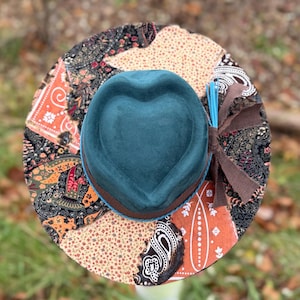 May include: A teal hat with a heart-shaped crown and a wide brim. The brim is decorated with a patchwork of paisley and floral patterns in shades of orange, black, and cream. A brown ribbon and blue sticks are tied to the side.