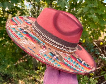 May include: A burgundy fedora-style hat with a wide brim. The brim features a colorful geometric pattern in shades of red, blue, and brown. The hat is adorned with a brown band and decorative trim.