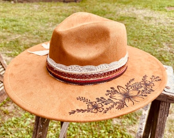 Puede incluir: Sombrero fedora marrón claro con ala ancha. El sombrero presenta una banda decorativa con detalles de encaje y cinta. Un diseño floral está grabado en el ala. Una pluma blanca está adherida al sombrero.