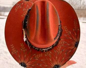 May include: A burnt orange cowboy hat with a decorative floral design. The hat features a dark brown brim and crown, with painted sunflowers and wheat stalks. The brim is adorned with black beading and feathers.