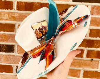 Pode incluir: Chapéu de cowboy de feltro branco com uma pena turquesa e marrom, uma borda de contas turquesa e marrom e um lenço estampado em vermelho, laranja e turquesa amarrado ao redor da coroa. O chapéu tem uma imagem vintage de um grupo de pessoas a cavalo.
