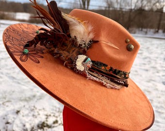 May include: An orange-colored hat with a wide brim, adorned with feathers, a decorative band, and a Native American-themed pin. The hat features a floral design on the brim and is embellished with small, round studs.