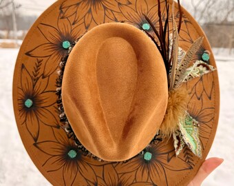 May include: A brown suede hat with a rounded crown and wide brim. The brim is decorated with black floral designs and turquoise-colored embellishments. The hat is adorned with feathers and a decorative fabric.
