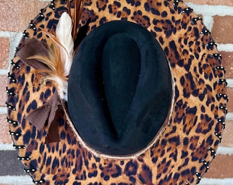 May include: A black fedora hat with a leopard print brim. The brim is decorated with black stitching and small white beads. The hat is adorned with brown and white feathers and a brown ribbon.