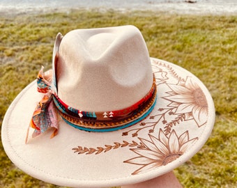 Puede incluir: Sombrero fedora beige con una banda decorativa con un patrón del suroeste y detalles turquesa. El ala del sombrero está adornada con un diseño de girasol, una bufanda colorida y una pluma. Un accesorio elegante.