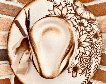 May include: A white felt cowboy hat with a brown floral design burned into the brim. The hat has a brown ribbon with a feather accent.