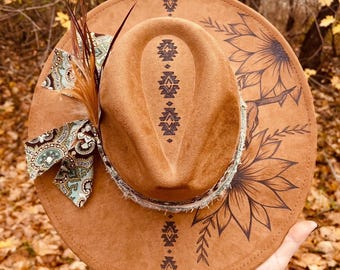 May include: A brown fedora-style hat with a wide brim, decorated with a sunflower design and tribal patterns. The hat features a decorative ribbon and feather accent. The hat is held by a person.